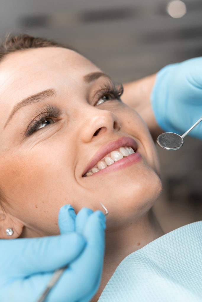 A Patient In Dental Chair Rejoices In Her New Smile. A Dentist, Looking Closely At Result, Provides High Quality Dental Care That Not Only Solves Problems, But Also Gives Confidence To The Patient.
